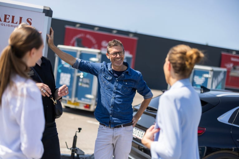 Smiling man talking to a group near branded Freetrailer units, illustrating partnership opportunities and how to become partner in a shared economy concept.
